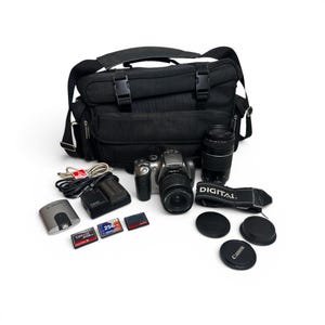 May include: A black camera bag containing a silver Canon digital SLR camera with a lens, a separate telephoto lens, a black camera strap with the word DIGITAL, three memory cards, a battery charger, a battery, a USB cable, and three lens caps.