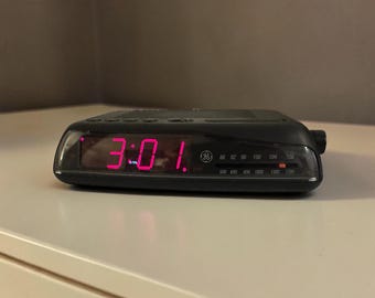 May include: A black GE digital alarm clock radio with a red LED display showing the time 3:01. The front panel features a radio frequency tuner with AM and FM scales, a GE logo, and a horizontal slider indicator for selecting radio stations.