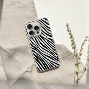 May include: A smartphone case with a black and white zebra stripe pattern. The case has a clear border and a camera cutout. The case is lying on a beige fabric with a sprig of dried flowers.