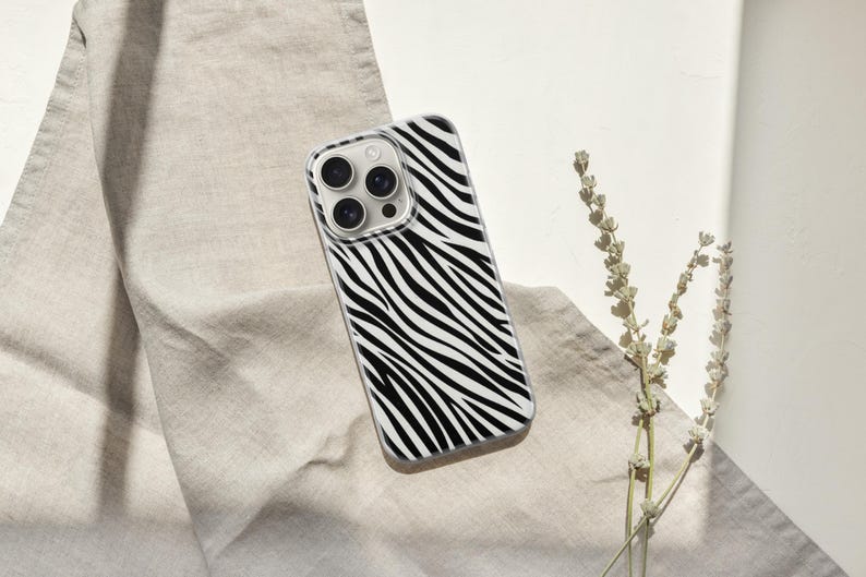 May include: A smartphone case with a black and white zebra stripe pattern. The case has a clear border and a camera cutout. The case is lying on a beige fabric with a sprig of dried flowers.