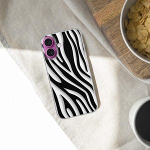 May include: A smartphone case with a black and white zebra stripe pattern. The case has a clear border and a pink camera lens protector. The phone case is lying on a white surface next to a bowl of cereal and a cup of coffee.
