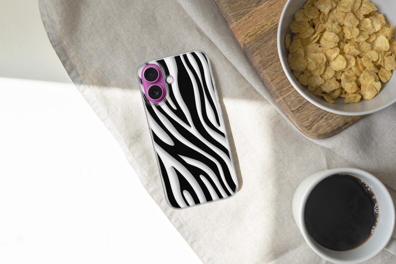 May include: A smartphone case with a black and white zebra stripe pattern. The case has a clear border and a pink camera lens protector. The phone case is lying on a white surface next to a bowl of cereal and a cup of coffee.