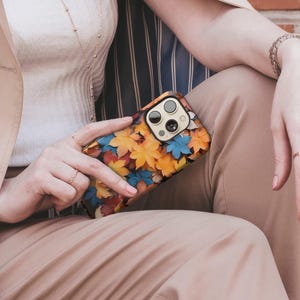 May include: A smartphone case featuring a colorful autumn leaf design in shades of yellow, orange, red, and blue. The case has a black border and is held by a person. The phone case is a protective accessory.