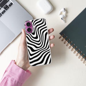 May include: A clear phone case with a black and white wavy pattern is held in a hand. The phone case has a purple camera lens protector. A laptop, earbuds, and a spiral-bound notebook are also visible.