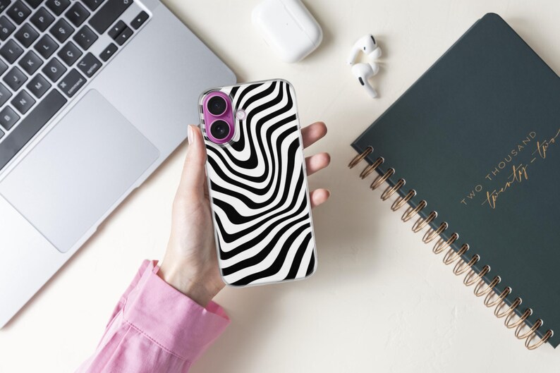 May include: A clear phone case with a black and white wavy pattern is held in a hand. The phone case has a purple camera lens protector. A laptop, earbuds, and a spiral-bound notebook are also visible.