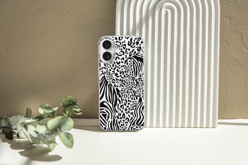 May include: A clear phone case featuring a black and white animal print design. The pattern combines zebra stripes, leopard spots, and other abstract shapes. The case is shown standing upright against a white, arched background.