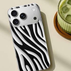 May include: A clear phone case with a black and white zebra stripe pattern. The case has a camera cutout with three lenses and a flash. The phone case is on a light beige surface.
