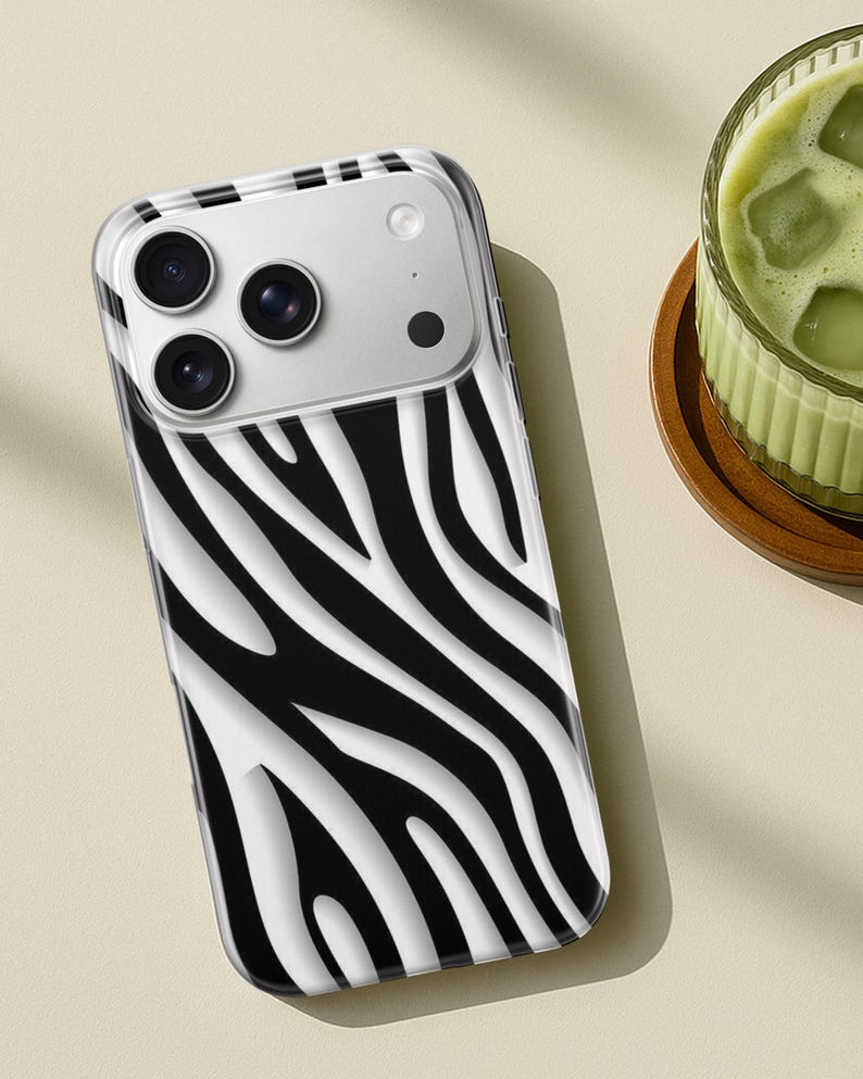 May include: A clear phone case with a black and white zebra stripe pattern. The case has a camera cutout with three lenses and a flash. The phone case is on a light beige surface.