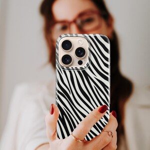 May include: A smartphone case with a black and white zebra stripe pattern. The case has a clear border and a camera module with multiple lenses. The phone is held up by a person with red nail polish.