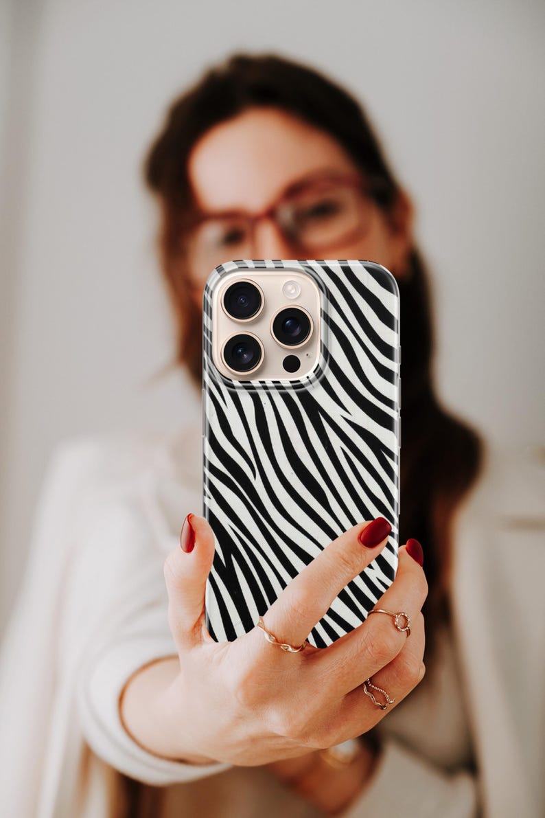May include: A smartphone case with a black and white zebra stripe pattern. The case has a clear border and a camera module with multiple lenses. The phone is held up by a person with red nail polish.
