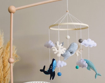 May include: A wooden baby mobile with a white, gray, and blue whale, a blue and white fish, and a white coral hanging from a wooden ring. The mobile is attached to a wooden stand with a white base.