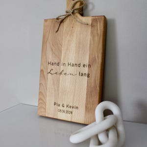 May include: A wooden cutting board with a handle and a rope tied around it. The board is engraved with the text "Hand in Hand ein Leben lang" and "Pia & Kevin 08.06.2024".