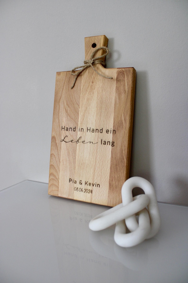 May include: A wooden cutting board with a handle and a rope tied around it. The board is engraved with the text "Hand in Hand ein Leben lang" and "Pia & Kevin 08.06.2024".