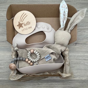 May include: A gift box filled with baby items, including a wooden sign that says "hello little one", a silicone bib, a muslin cloth, a wooden teether, and a bunny rattle.