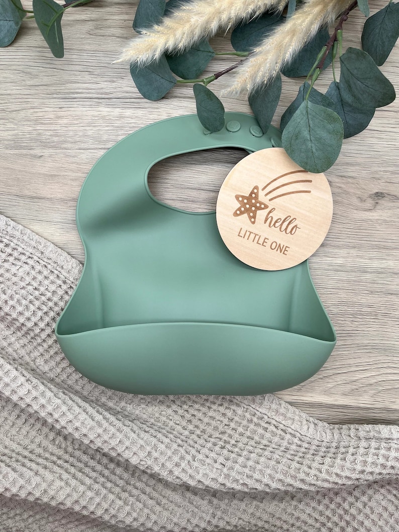 May include: A green silicone baby bib with a wooden tag that says "hello little one" with a starfish and shooting star design.