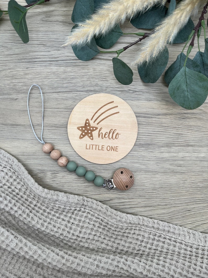 May include: A wooden baby announcement disc with a star and shooting star design. The disc says "hello little one". A wooden and silicone teething toy with a green silicone bead chain is also pictured.