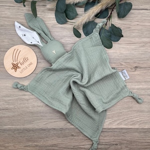 May include: A sage green muslin baby security blanket with a knotted corner and a bunny head attached. The bunny has white ears with black stars and a white triangle on its chest. The blanket has a tag that says "hello little humans".