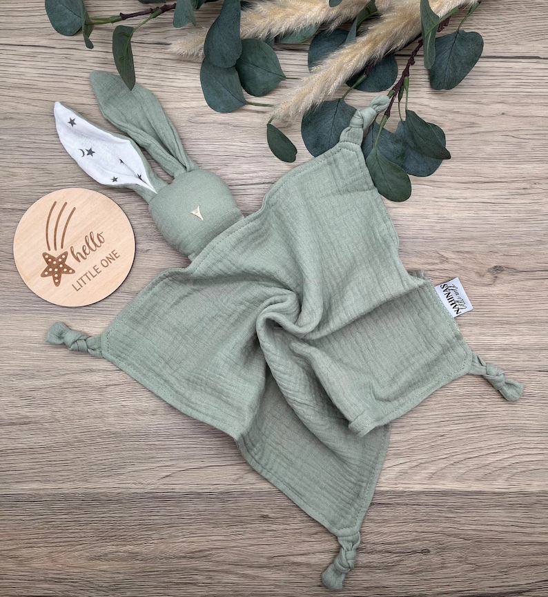 May include: A sage green muslin baby security blanket with a knotted corner and a bunny head attached. The bunny has white ears with black stars and a white triangle on its chest. The blanket has a tag that says "hello little humans".