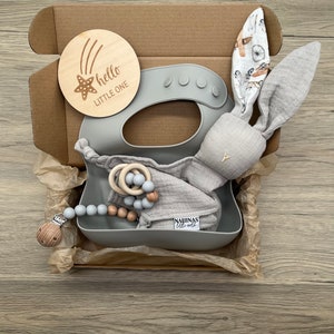 May include: A gray gift box filled with baby items. The box contains a gray silicone bib, a gray bunny rattle, a wooden teether, and a gray and wood bead necklace. The box also includes a wooden sign that says "hello little one" with a star and shooting stars.