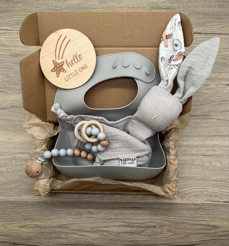 May include: A gray gift box filled with baby items. The box contains a gray silicone bib, a gray bunny rattle, a wooden teether, and a gray and wood bead necklace. The box also includes a wooden sign that says "hello little one" with a star and shooting stars.