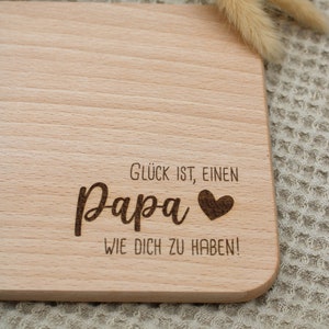 Może przedstawiać: Drewniana deska do krojenia z tekstem "Gl&uuml;ck ist, einen Papa wie dich zu haben!" (Szczęście to mieć takiego tatę jak ty!) i małym sercem.