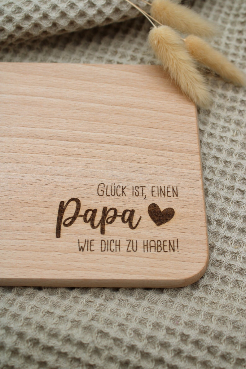 Może przedstawiać: Drewniana deska do krojenia z tekstem "Gl&uuml;ck ist, einen Papa wie dich zu haben!" (Szczęście to mieć takiego tatę jak ty!) i małym sercem.