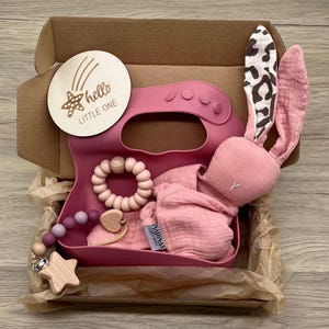 May include: A pink gift box filled with baby items, including a pink bunny rattle, a pink silicone bib, a wooden teether, and a wooden star rattle. The box also includes a wooden sign that says "hello little one".