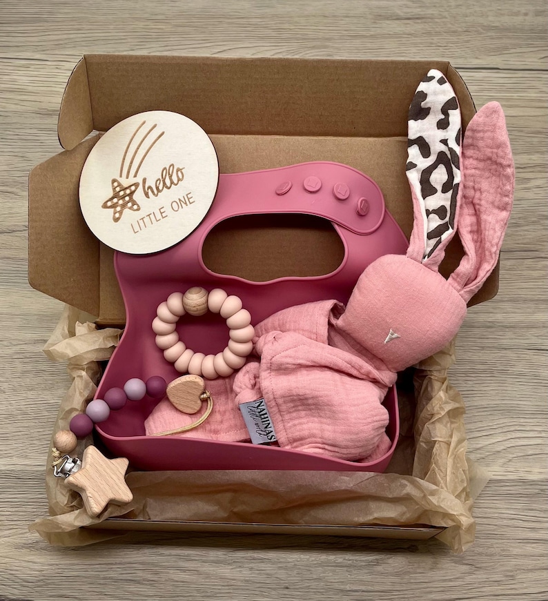 May include: A pink gift box filled with baby items, including a pink bunny rattle, a pink silicone bib, a wooden teether, and a wooden star rattle. The box also includes a wooden sign that says "hello little one".
