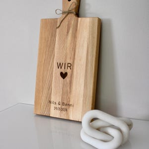 May include: A wooden cutting board with a handle and the words "WIR" and a heart, followed by "Nils & Benni 24.05.2024" engraved on the surface.