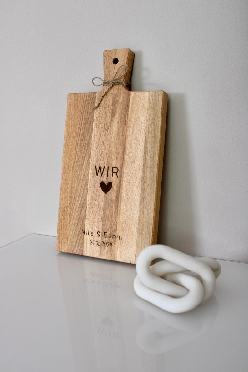 May include: A wooden cutting board with a handle and the words "WIR" and a heart, followed by "Nils & Benni 24.05.2024" engraved on the surface.