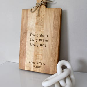 May include: A wooden cutting board with a handle and a rope tied around it. The board is engraved with the text "Ewig dein, Ewig mein, Ewig uns, Anna & Tom, 25.05.2019".