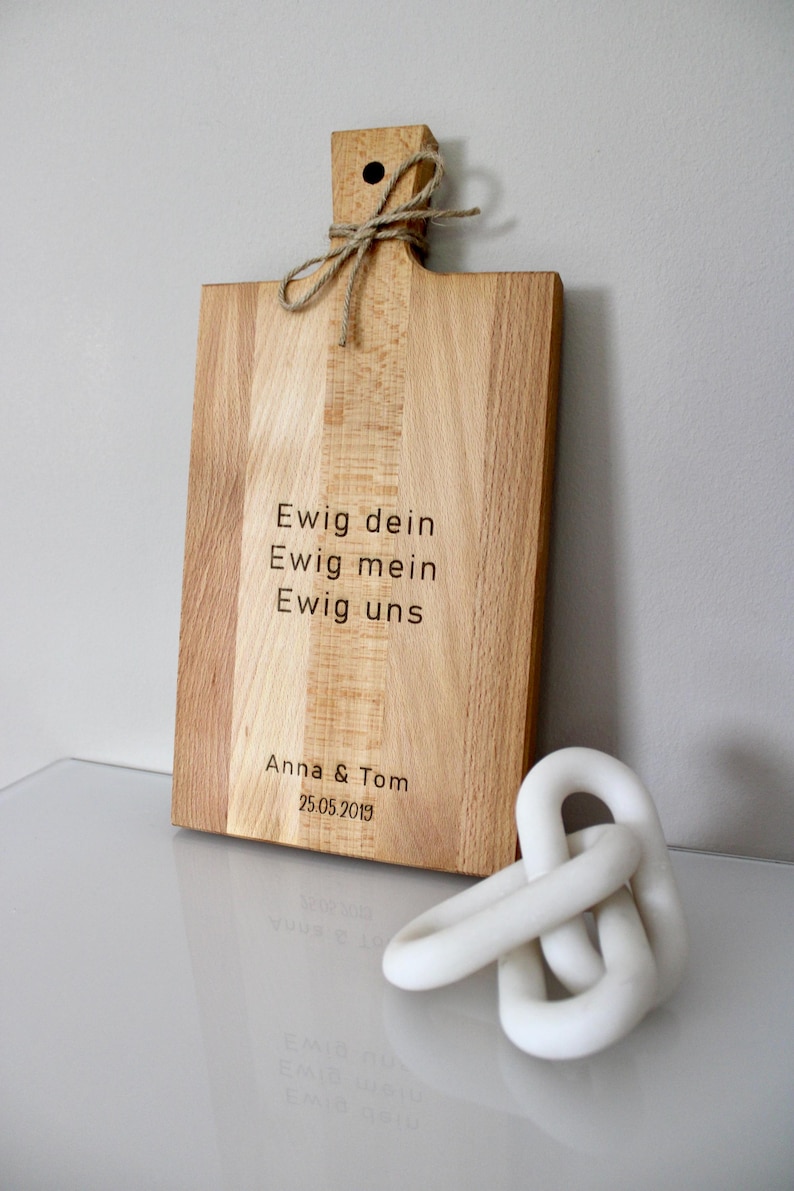 May include: A wooden cutting board with a handle and a rope tied around it. The board is engraved with the text "Ewig dein, Ewig mein, Ewig uns, Anna & Tom, 25.05.2019".