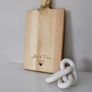 May include: A personalized wooden cutting board with the text "Alice & Florian 31.08.2024" and a small heart engraved on the surface. The board has a handle and is made of light wood.