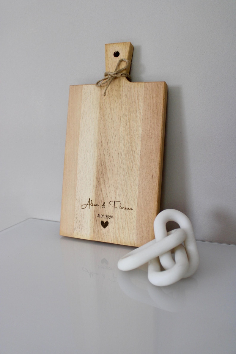 May include: A personalized wooden cutting board with the text "Alice & Florian 31.08.2024" and a small heart engraved on the surface. The board has a handle and is made of light wood.