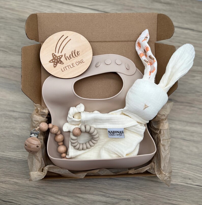 May include: A gift box filled with baby items, including a wooden engraved sign that says "hello little one", a beige silicone bib, a white muslin cloth with the text "Nahinas little ones", a beige silicone teether, a wooden bead necklace with a clip, and a white bunny plush toy.