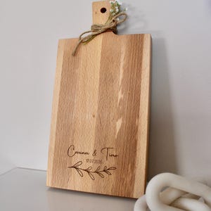 May include: A wooden cutting board with a natural finish and a handle. The board is engraved with the text "Carina & Timo 17.07.2020" and a floral design.
