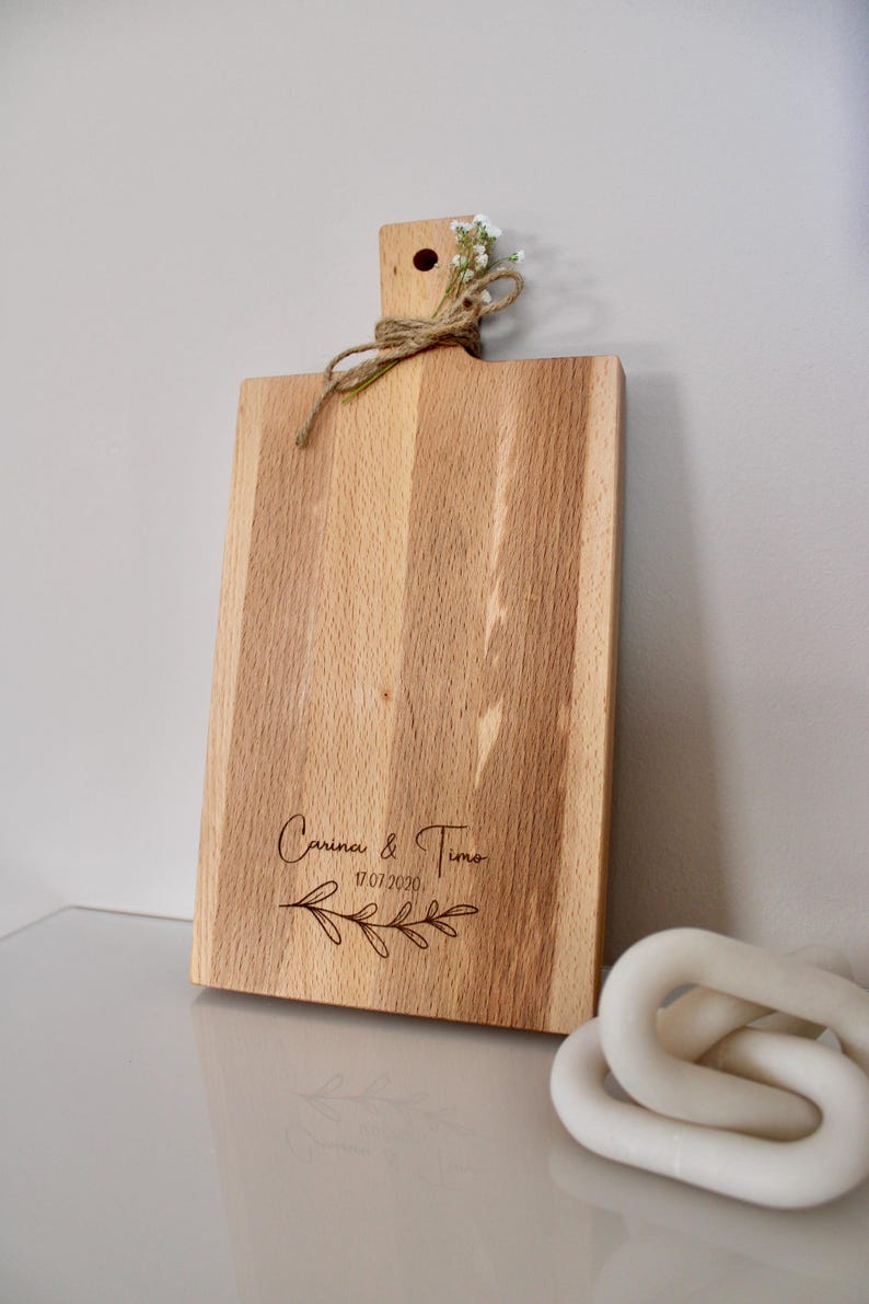 May include: A wooden cutting board with a natural finish and a handle. The board is engraved with the text "Carina & Timo 17.07.2020" and a floral design.