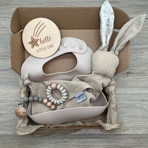 May include: A gift box filled with baby items, including a wooden sign that says "hello little one", a beige bunny plush toy, a silicone bib, a muslin cloth, a wooden and grey bead teether, and a wooden and grey bead clip.