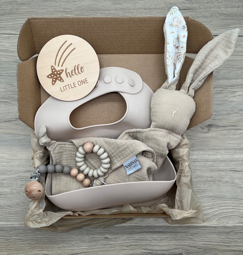 May include: A gift box filled with baby items, including a wooden sign that says "hello little one", a beige bunny plush toy, a silicone bib, a muslin cloth, a wooden and grey bead teether, and a wooden and grey bead clip.