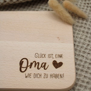 Może przedstawiać: Drewniana deska do krojenia z wygrawerowanym tekstem "Gl&uuml;ck ist, eine Oma &hearts; wie dich zu haben!" Deska leży na beżowej, teksturowanej powierzchni.
