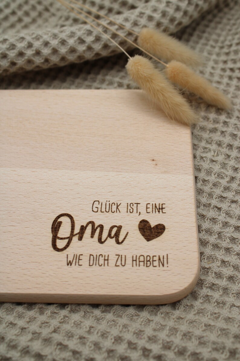 Może przedstawiać: Drewniana deska do krojenia z wygrawerowanym tekstem "Gl&uuml;ck ist, eine Oma &hearts; wie dich zu haben!" Deska leży na beżowej, teksturowanej powierzchni.
