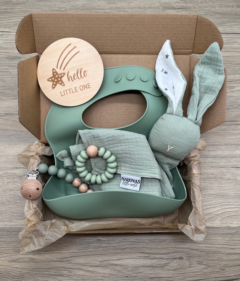 May include: A green gift box filled with baby items. The box contains a wooden sign that says "hello little one", a green silicone bib, a green muslin cloth, a wooden teether, and a green bunny rattle.