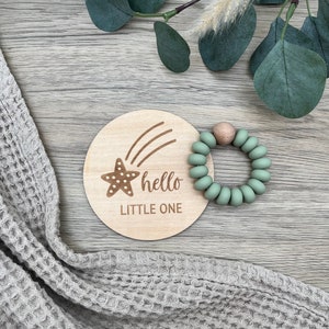 May include: A wooden baby milestone disc with a starfish and the text "hello little one". A green silicone teething ring is next to the disc.