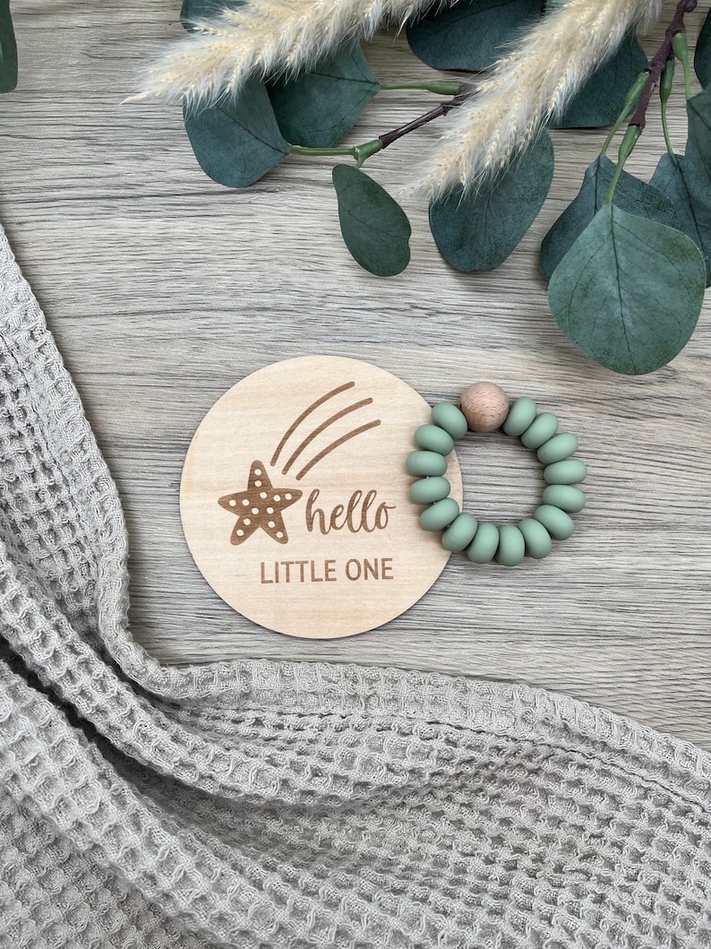 May include: A wooden baby milestone disc with a starfish and the text "hello little one". A green silicone teething ring is next to the disc.