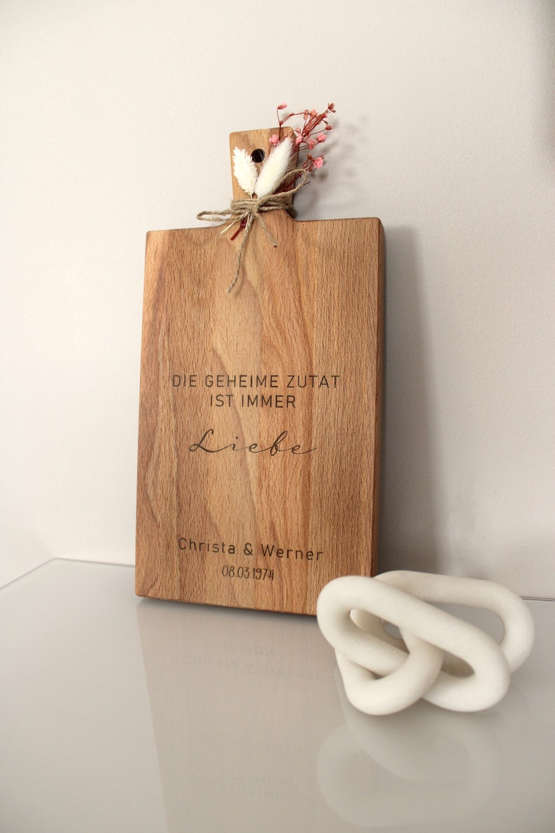 May include: A wooden cutting board with the text "DIE GEHEIME ZUTAT IST IMMER Liebe" and "Christa & Werner 08.03.1974" engraved on it. The board is decorated with dried flowers and twine.