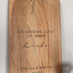 May include: A wooden cutting board with the text "Die geheime Zutat ist immer Liebe" and "Christa & Werner 08.03.1974" engraved on it. The board is decorated with dried flowers.