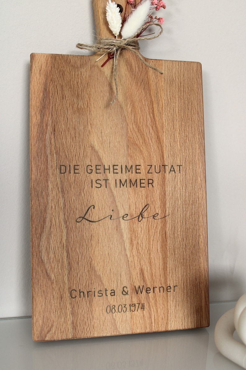 May include: A wooden cutting board with the text "Die geheime Zutat ist immer Liebe" and "Christa & Werner 08.03.1974" engraved on it. The board is decorated with dried flowers.