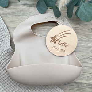 May include: A light beige silicone bib with a pocket for catching food. A wooden circle with a brown star and the text "hello little one" is placed on top of the bib.
