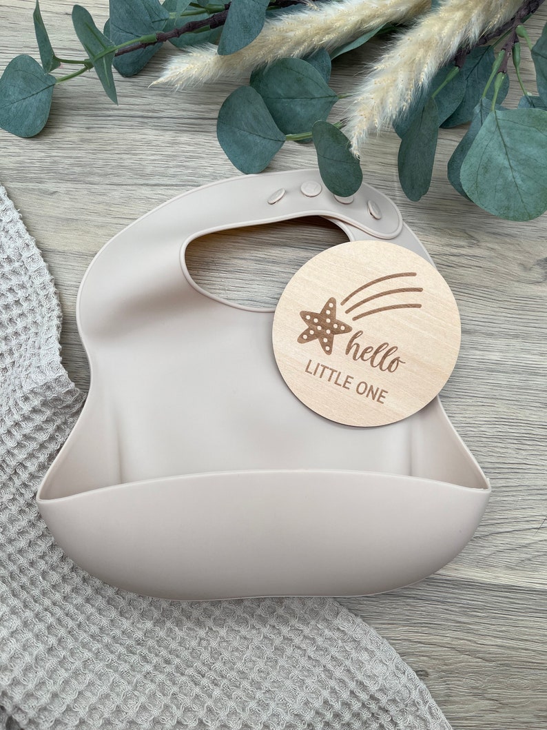 May include: A light beige silicone bib with a pocket for catching food. A wooden circle with a brown star and the text "hello little one" is placed on top of the bib.