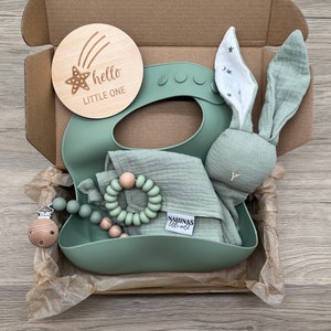 May include: A green gift box filled with baby items. The box contains a wooden sign that says "hello little one", a green silicone bib, a green muslin cloth, a wooden teether, and a green bunny rattle.